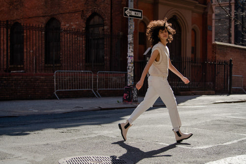 A person with curly hair wearing a sleeveless top, white pants, and boots confidently walks across a street in a city, passing a One Way sign and a brick building in the background.