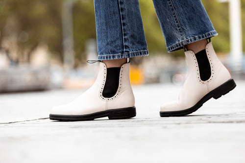 Person wearing light-colored Chelsea boots and blue jeans, standing on a sidewalk outdoors with a blurred background of trees and street elements.