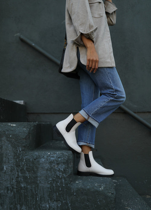 A person wearing blue jeans, a beige oversized shirt, and white Chelsea boots stands on dark concrete steps against a gray wall. Their face is not visible.
