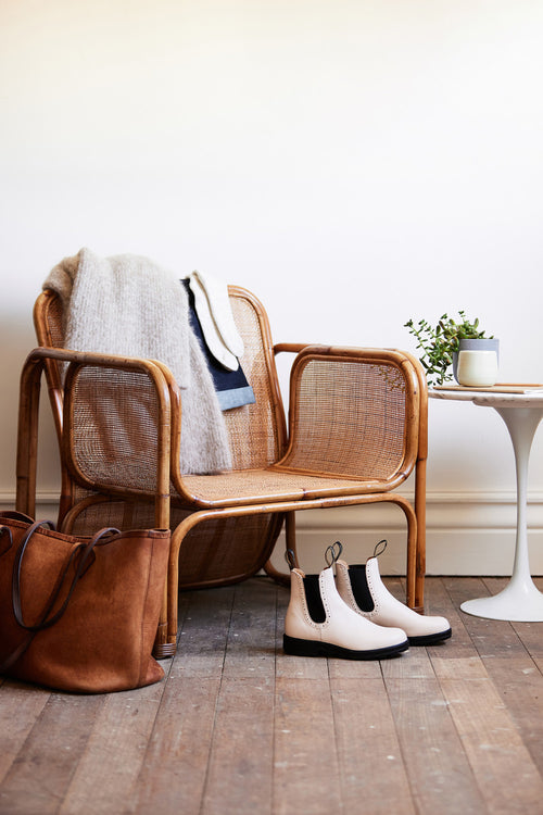 A cozy scene featuring a woven rattan chair draped with a soft gray blanket, white boots on the wooden floor, a brown leather bag nearby, and a small white table holding a plant and a mug.