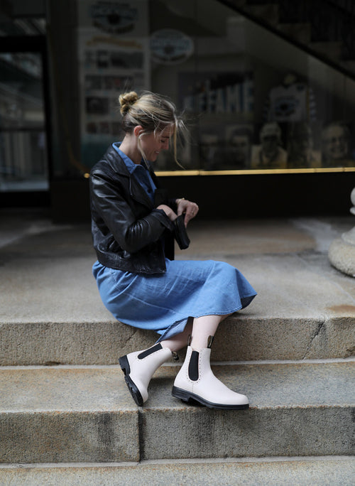 A woman in a blue dress, black leather jacket, and white ankle boots sits on outdoor stone steps, looking down while adjusting something in her hands.