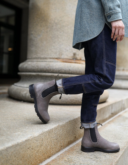 A person in dark rolled-up jeans, gray Chelsea boots, and a textured gray jacket stands on stone steps next to a large column, lifting one leg slightly as if about to walk.