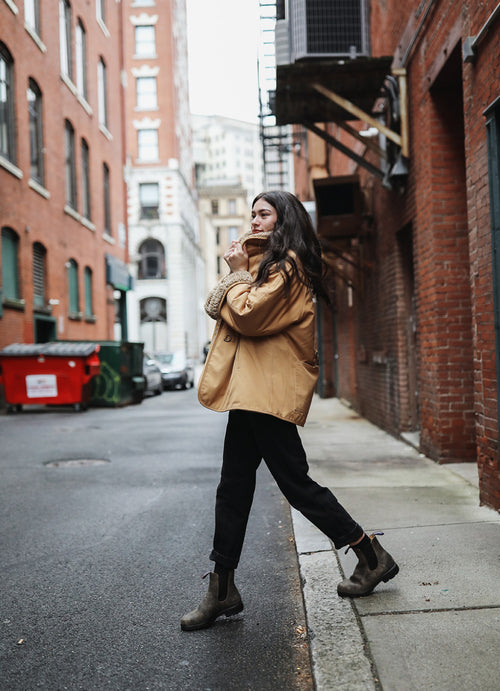 A woman wearing a tan coat, black pants, and boots walks along a city alley lined with red brick buildings. She looks over her shoulder and smiles, with one hand raised near her face.