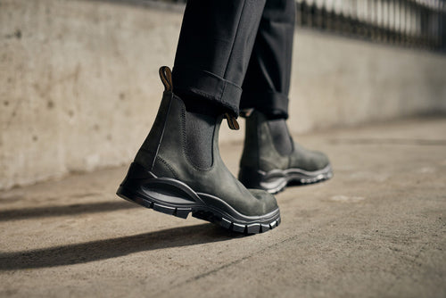 Close-up of a person wearing black pants and dark green Chelsea boots with thick, rugged soles, walking on a concrete surface near a blurred fence.