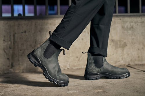 A person wearing black pants and rugged dark gray Chelsea boots stands on a concrete surface, with a metal railing blurred in the background.
