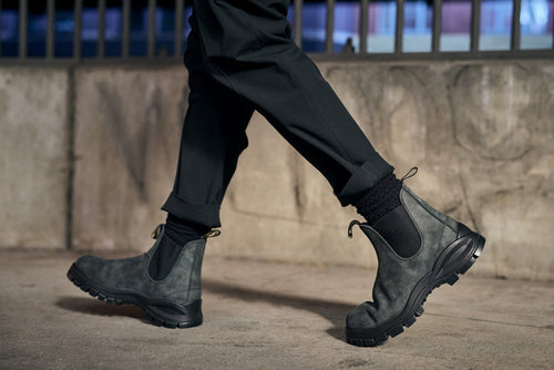 A person wearing black pants, black socks, and rugged gray Chelsea boots is walking on a concrete surface near a wall with metal bars in the background.