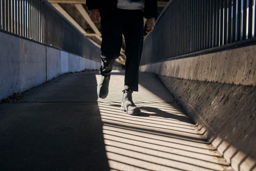 A person wearing black boots and dark pants walks on a sunlit, shadow-striped pedestrian bridge with metal railings and concrete walls.