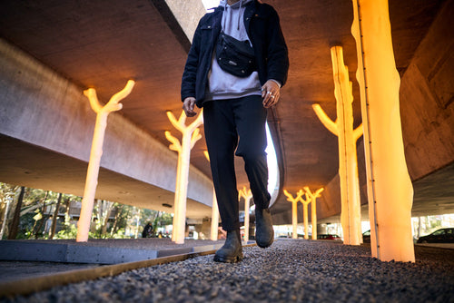 A person walks on a gravel path under a bridge with glowing, tree-like light installations. The photo is taken from a low angle, focusing on the persons legs and feet.
