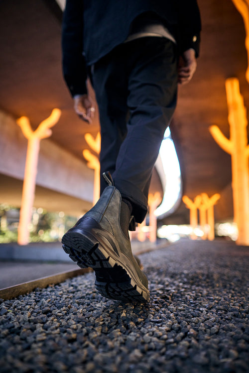 A person wearing black boots and dark pants walks on gravel under an overpass with orange, tree-shaped lights in the background. The perspective is low, focused on the sole of one boot.