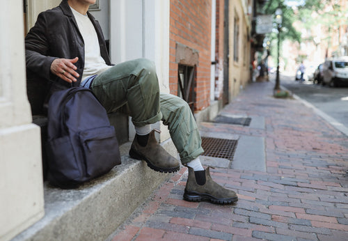 A person wearing green cargo pants, brown boots, and a dark jacket sits on a stone step holding a dark backpack on a brick sidewalk in an urban setting.