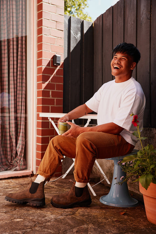 A smiling man in a white t-shirt, brown pants, and boots sits outdoors on a blue stool, holding a green mug. He is next to a small white table, with plants and a brick wall in the background.