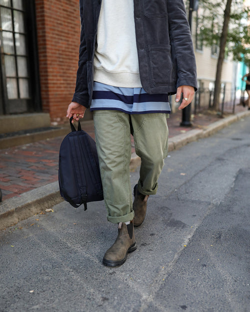 Person walking on a street, wearing cuffed olive green pants, brown boots, a striped shirt under a light sweater, and a dark jacket, holding a black backpack by their side. Brick building and trees in the background.