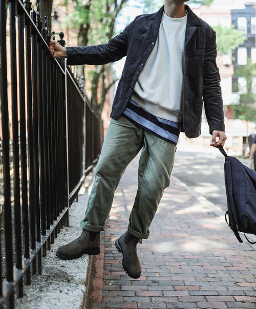 A person in a casual outfit stands on a brick sidewalk, holding a navy backpack and leaning on a black iron fence. They wear a dark jacket, white shirt, green pants, and brown boots. Trees and buildings are in the background.