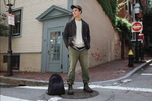 A young man in casual clothing stands on a street corner, smiling with hands in pockets. He wears a cap, jacket, green pants, and boots. A black backpack sits on the ground nearby. Brick buildings and street signs are visible.