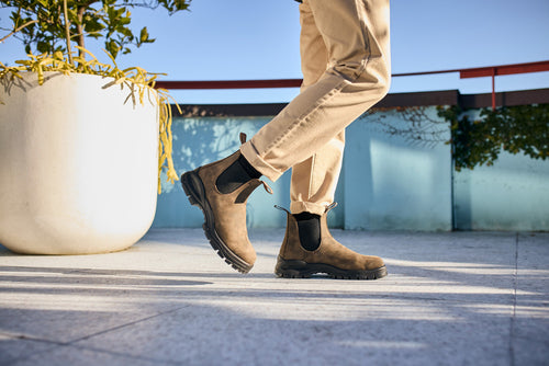 A person wearing tan pants and brown Chelsea boots walks outdoors on a sunlit patio with a large white planter and a blue wall in the background.