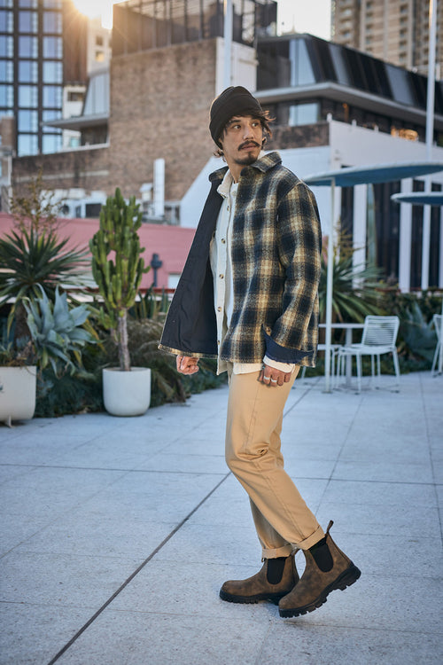 A man wearing a black beanie, plaid jacket, light shirt, khaki pants, and brown boots stands on a modern outdoor patio with plants and city buildings in the background.