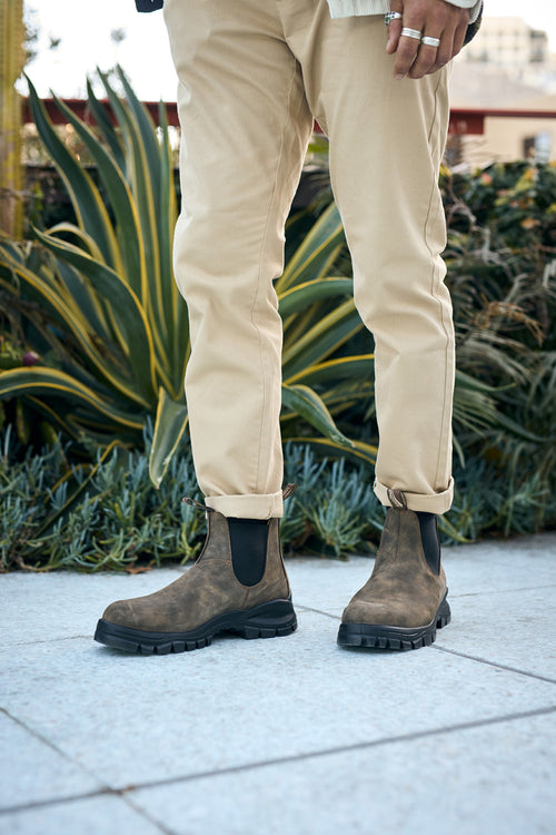 A person wearing tan pants and brown Chelsea boots stands on a sidewalk in front of green, leafy plants. Only the lower half of the body is visible.