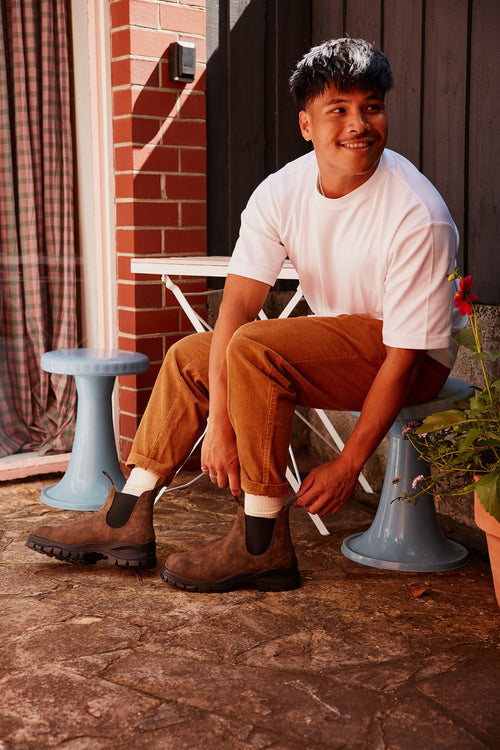 A man in a white t-shirt and brown pants sits on a blue stool outdoors, smiling while putting on brown boots. He is on a stone patio beside a small table, with a potted plant nearby.