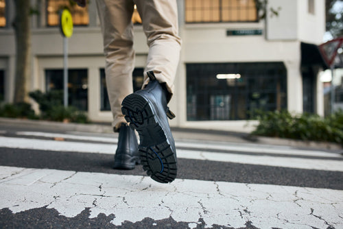 A person wearing tan pants and black boots is walking across a crosswalk on a city street. The photo is taken from behind, focusing on the sole of one boot in mid-step.