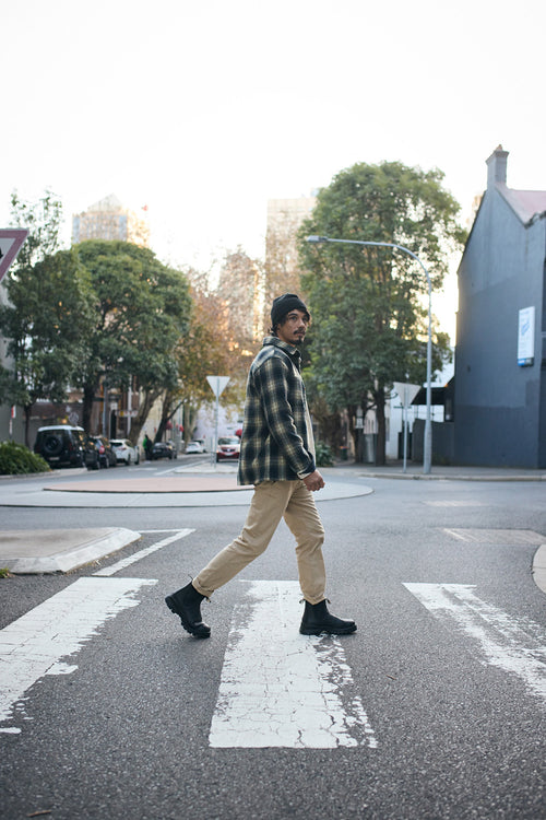A man wearing a plaid shirt, beige pants, and black boots walks across a crosswalk on a city street, surrounded by trees and buildings.
