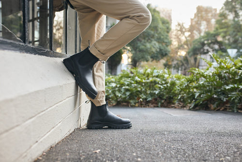 Person in tan pants wearing black boots leans against a white wall outdoors on a sidewalk, with greenery and trees in the background.