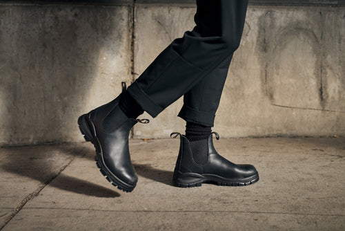 A person wearing black ankle boots, black socks, and dark pants walks on a concrete sidewalk in front of a weathered, gray concrete wall.