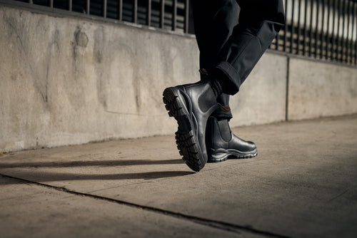 A person wearing black, rugged boots and black pants walks on a concrete sidewalk next to a weathered wall with metal bars. The scene is outdoors and the lighting is moody.