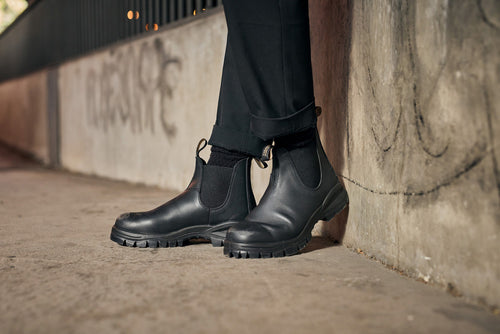 A person wearing black pants and black Chelsea boots stands on a concrete sidewalk next to a graffiti-covered wall. The photo focuses on the boots and lower legs.