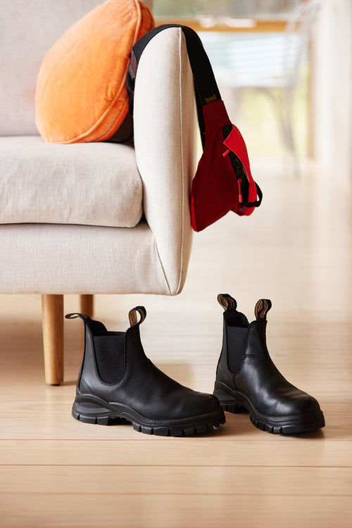 A pair of black Chelsea boots sits on a light wooden floor next to a beige sofa with an orange cushion. A red bag hangs over the sofa’s armrest.