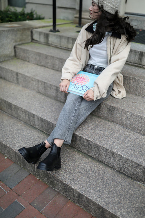 A woman in a beige jacket, gray jeans, and black boots sits on outdoor stone steps, holding a book with a colorful flower illustration on the cover. She is looking to the side and wearing a knit beanie.