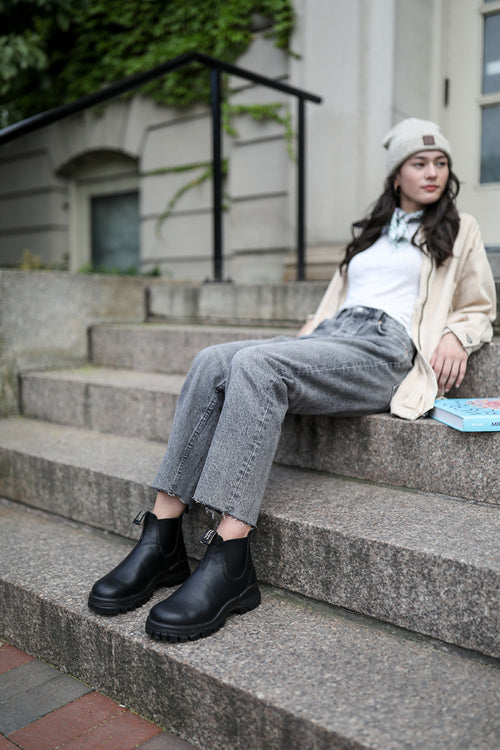 A young woman in a beanie, light jacket, white shirt, and frayed gray jeans sits on outdoor stone steps, wearing black boots. A closed book lies beside her, and greenery is visible in the background.