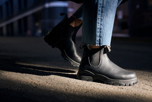 Close-up of a person wearing black leather Chelsea boots and blue jeans, standing on a sunlit pavement with shadows and blurred urban buildings in the background.