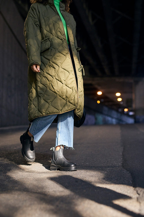 A person wearing a long green quilted coat, blue jeans, and black boots walks on a shaded urban street with lights illuminating the background.