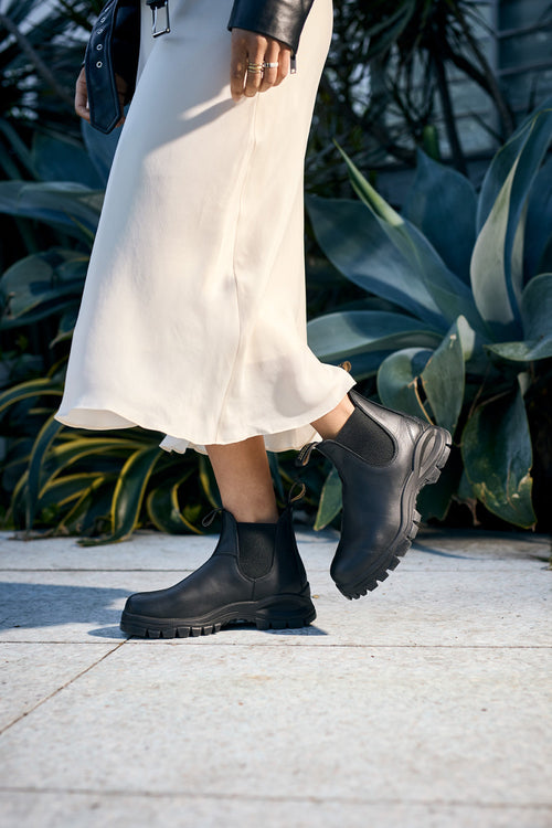 A person wearing a cream-colored skirt and black ankle boots walks on a stone path, with large green plants visible in the background.