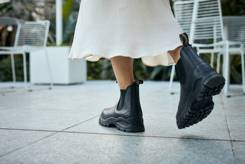 A person wearing a flowing white skirt and black ankle boots walks on a tiled outdoor surface, with white metal chairs and greenery in the background.