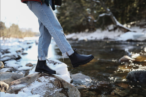 A person wearing cuffed jeans and black boots balances on a rock beside a shallow, snow-lined river in a forested area during winter.