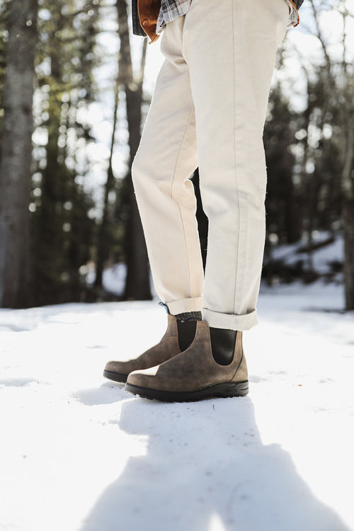 A person wearing light-colored pants and brown Chelsea boots stands on snow in a sunlit forest, with trees and shadows in the background.