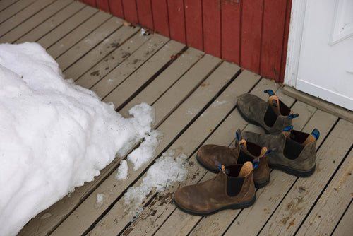 Three pairs of brown boots sit on a wooden porch next to a pile of melting snow, in front of a red-paneled building and a white door.