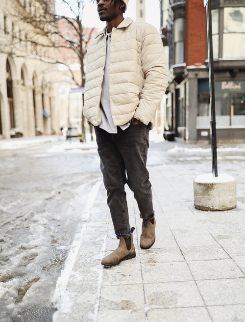 A person wearing a light beige puffer jacket, dark jeans, and brown boots stands on a snowy city sidewalk. The image is cropped above the mouth, showing only the lower face and body.