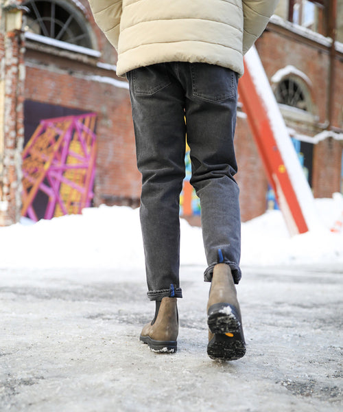 Person wearing a light jacket, dark jeans, and brown boots walks on a snowy sidewalk near a brick building with graffiti and colorful art structures in the background.