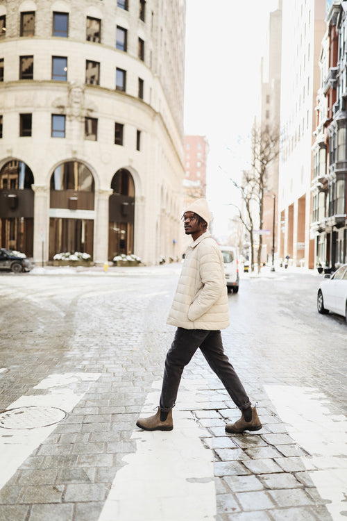 A man wearing a light jacket and beanie walks across a city street at a crosswalk on a bright, sunny day. Tall buildings and a few cars line the street in the background.