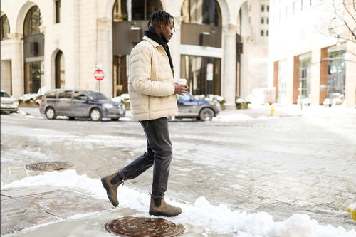 A man wearing a light-colored puffer jacket, black jeans, and brown boots walks across a snowy city street while holding a coffee cup. Cars and buildings are visible in the background.