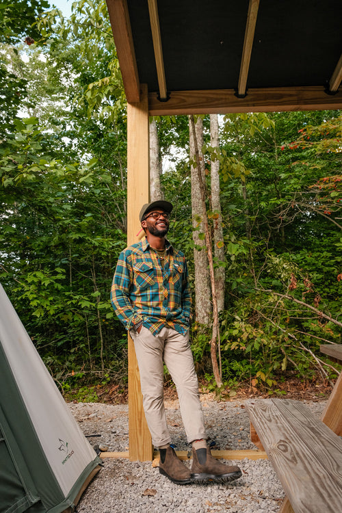 A man in a plaid shirt, light pants, boots, and a hat stands and smiles under a wooden shelter near a tent in a wooded outdoor area, with trees and greenery in the background.