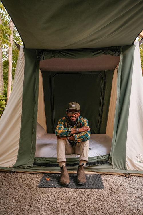 A man wearing glasses, a green hat, a plaid shirt, and brown boots sits smiling on the edge of a bed inside a canvas tent with the door open, surrounded by trees and gravel ground.