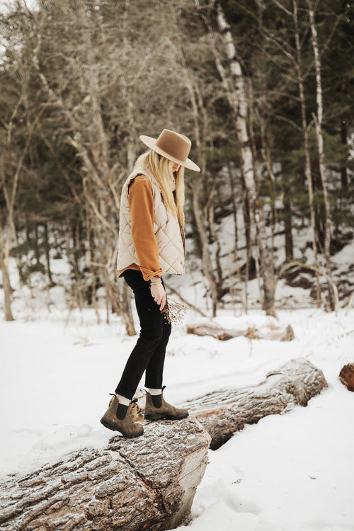 A person wearing a wide-brimmed hat, tan vest, brown sweater, black pants, and boots stands on a fallen log in a snowy forest, looking down and holding a small bunch of dried flowers.