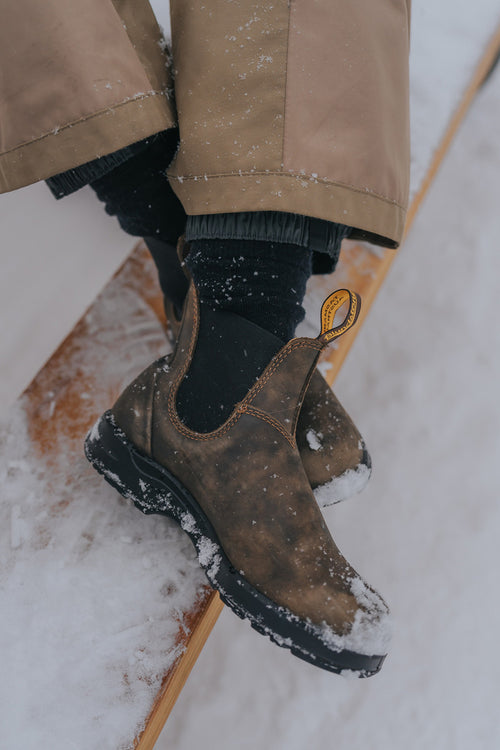 A person wearing brown leather boots, black socks, and tan pants sits on a snowy wooden surface. Snow is scattered on the boots and the wooden plank.
