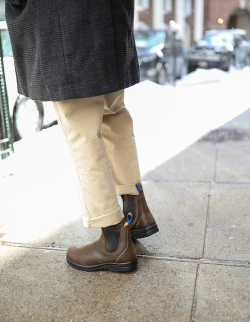 A person stands on a city sidewalk wearing a dark coat, beige pants, and brown Chelsea boots with blue heel tabs. Parked cars and blurred buildings are visible in the background.