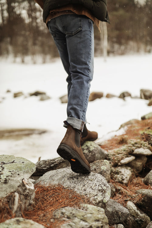 A person in jeans and brown boots steps across rocky ground outdoors, with snow and trees in the background. The photo is taken from behind, focusing on their legs and boots.