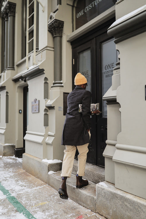 A person wearing a yellow beanie, dark coat, light pants, and boots steps up to enter a building with double doors on a snowy sidewalk.