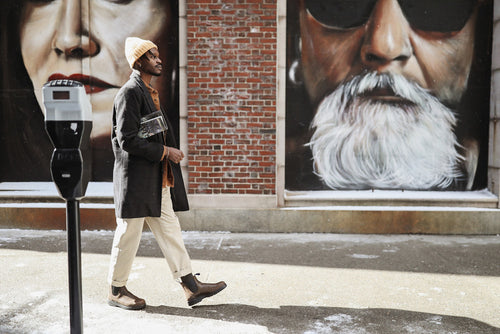 A man wearing a coat, beanie, and boots walks past a brick wall with large murals of faces, one of a woman and one of a man with a white beard and sunglasses, on a snowy sidewalk.